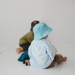 Two toddlers sitting on a white floor in a minimalist studio setting. The child at the front is wearing a light blue daycare bucket hat and a pale blue shirt, while the child behind wears an olive green shirt and a dark brown daycare bucket hat.