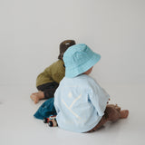 Two toddlers sitting on a white floor in a minimalist studio setting. The child at the front is wearing a light blue daycare bucket hat and a pale blue shirt, while the child behind wears an olive green shirt and a dark brown daycare bucket hat.