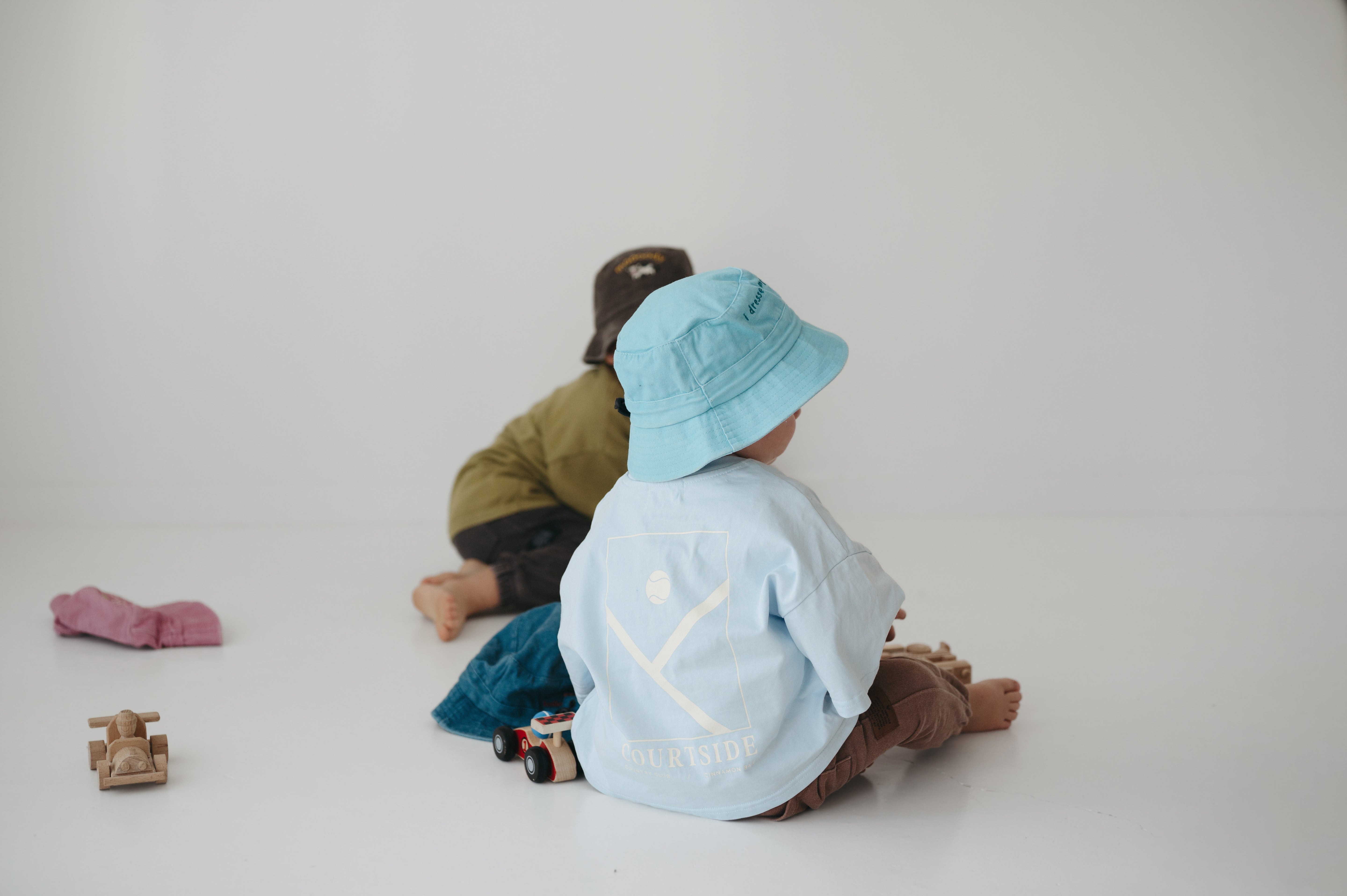 Two toddlers sitting on a white floor in a minimalist studio setting. The child at the front is wearing a light blue daycare bucket hat and a pale blue shirt, while the child behind wears an olive green shirt and a dark brown daycare bucket hat.