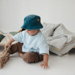 A toddler wearing a blue daycare washed cotton bucket hat, light blue t-shirt, and brown pants sits on the floor next to a daycare stacker bed, playing with a wooden fire truck. A blue gingham and sage green daycare bedding nap mats are displayed over the stacker bed in a bright, minimalist room.