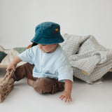 A toddler wearing a blue daycare washed cotton bucket hat, light blue t-shirt, and brown pants sits on the floor next to a daycare stacker bed, playing with a wooden fire truck. A blue gingham and sage green daycare bedding nap mats are displayed over the stacker bed in a bright, minimalist room.