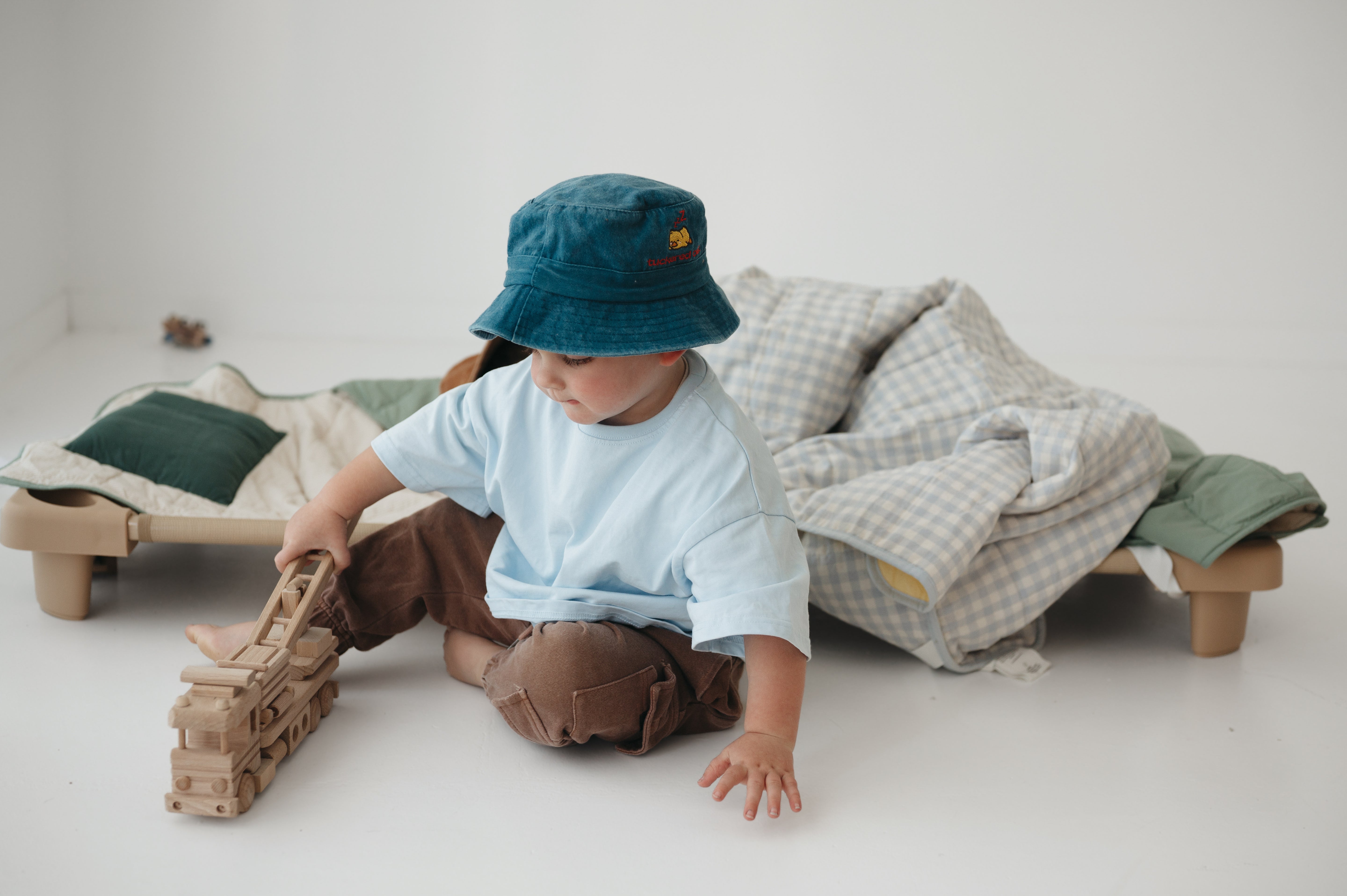 A toddler wearing a blue daycare washed cotton bucket hat, light blue t-shirt, and brown pants sits on the floor next to a daycare stacker bed, playing with a wooden fire truck. A blue gingham and sage green daycare bedding nap mats are displayed over the stacker bed in a bright, minimalist room.