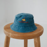 A dark blue washed cotton toddler daycare bucket hat with "tuckered out" embroidered in a red text on the front and baby yellow duck sleeping, displayed on a round wooden stool against a white background.