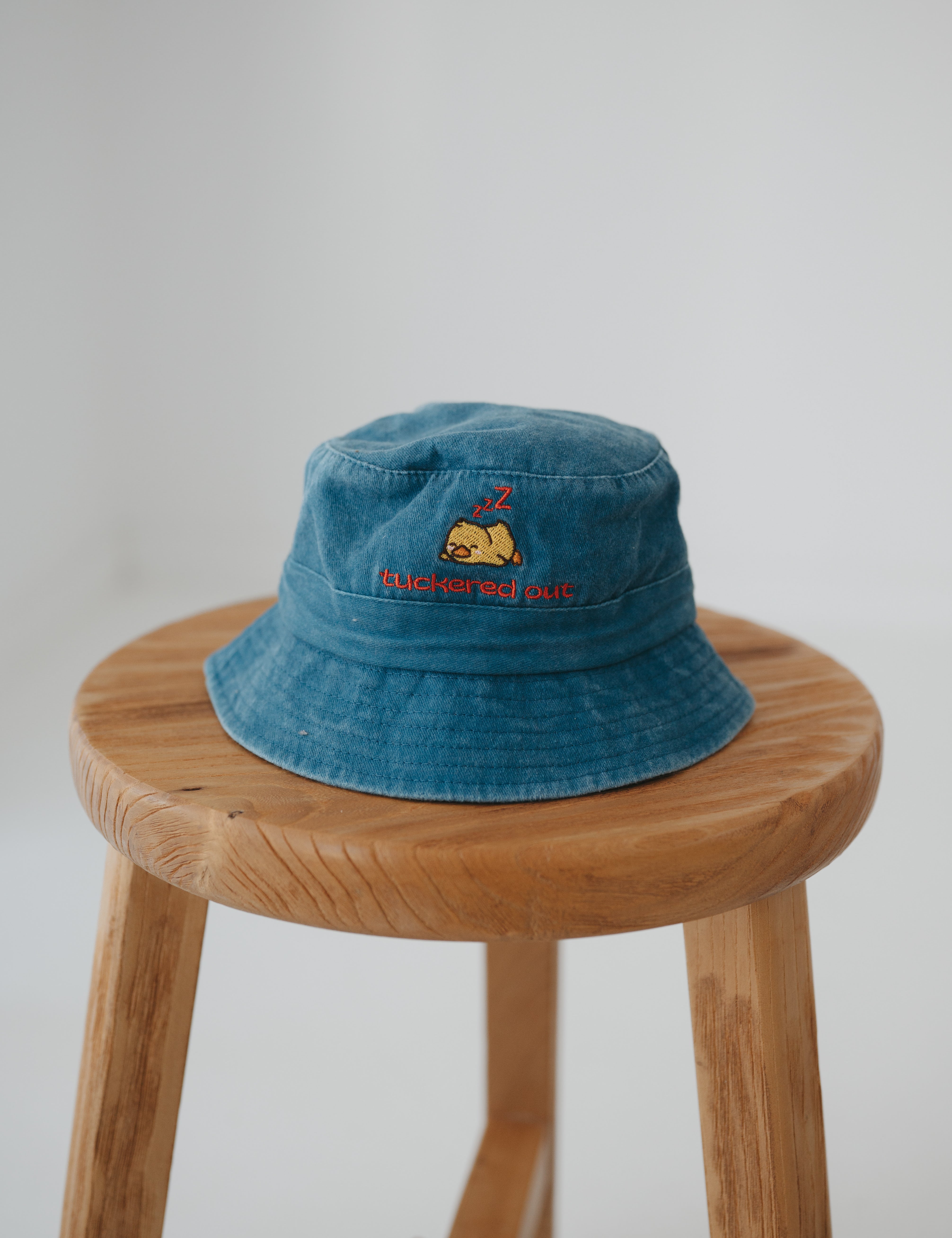 A dark blue washed cotton toddler daycare bucket hat with "tuckered out" embroidered in a red text on the front and baby yellow duck sleeping, displayed on a round wooden stool against a white background.