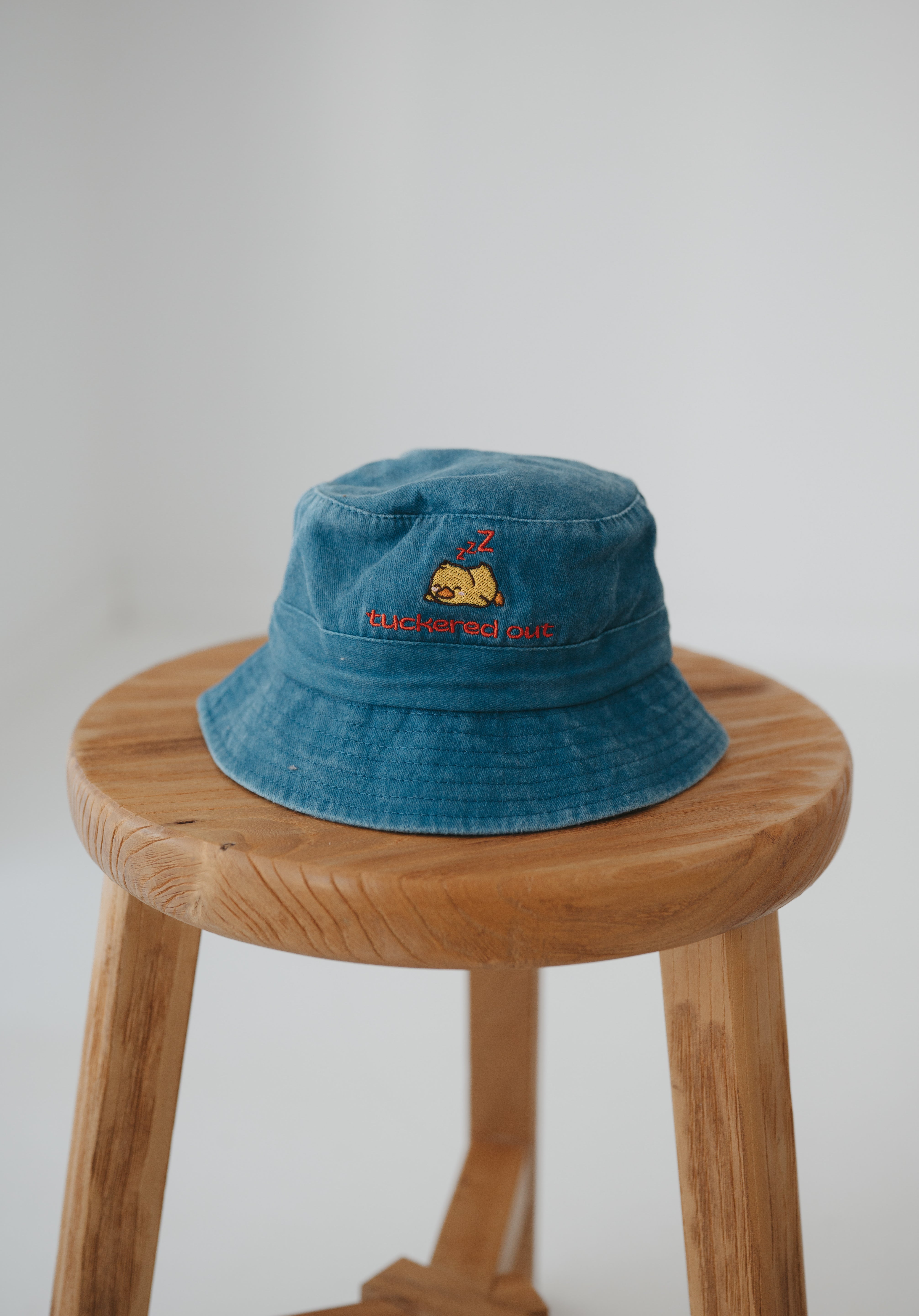 A dark blue washed cotton toddler daycare bucket hat with "tuckered out" embroidered in a red text on the front and baby yellow duck sleeping, displayed on a round wooden stool against a white background.