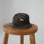 A brown washed cotton daycare bucket hat with "moooody" embroidered in yellow with an embroidered cow, displayed on a wooden stool against a light grey background.