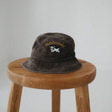 A brown washed cotton daycare bucket hat with "moooody" embroidered in yellow with an embroidered cow, displayed on a wooden stool against a light grey background.