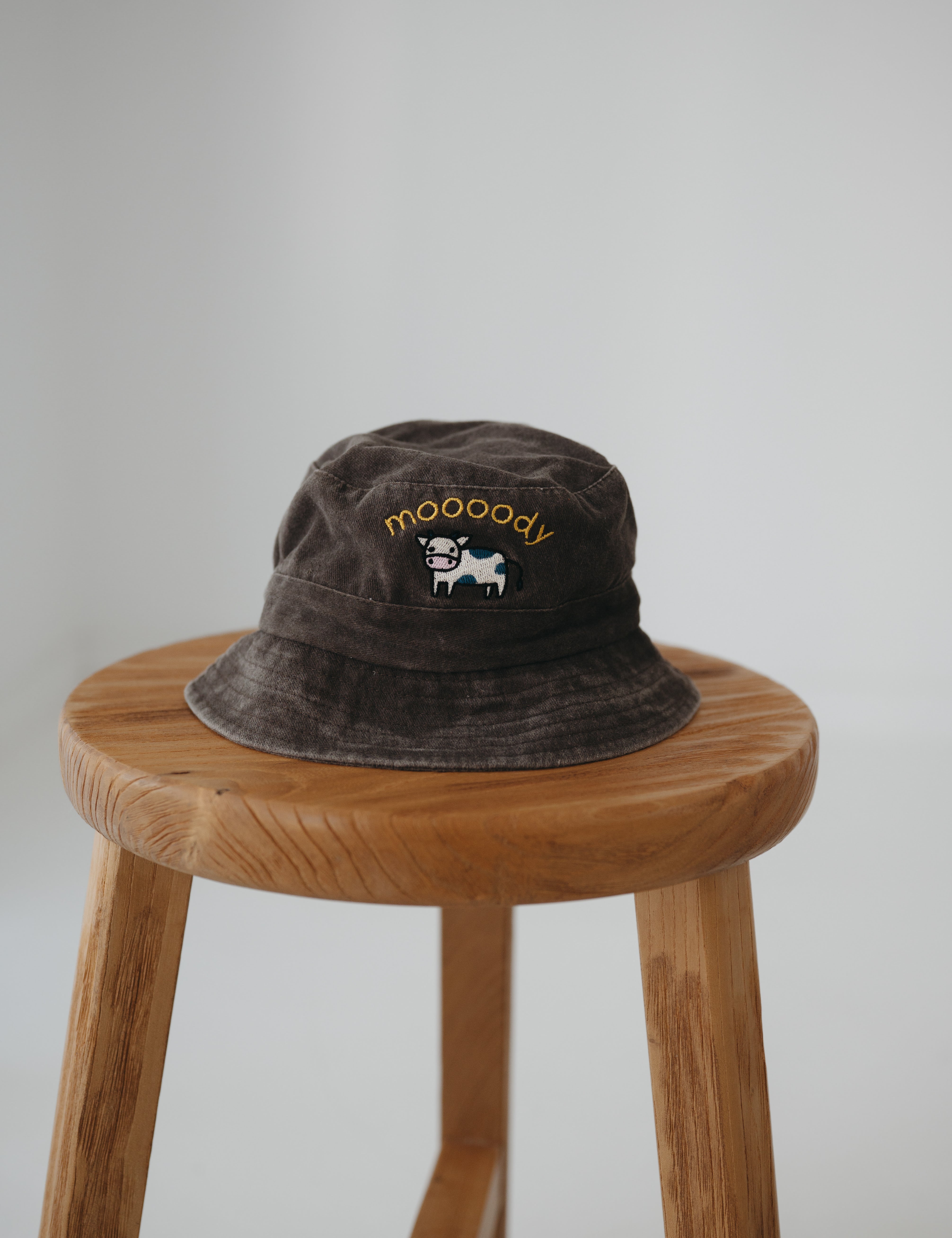 A brown washed cotton daycare bucket hat with "moooody" embroidered in yellow with an embroidered cow, displayed on a wooden stool against a light grey background.