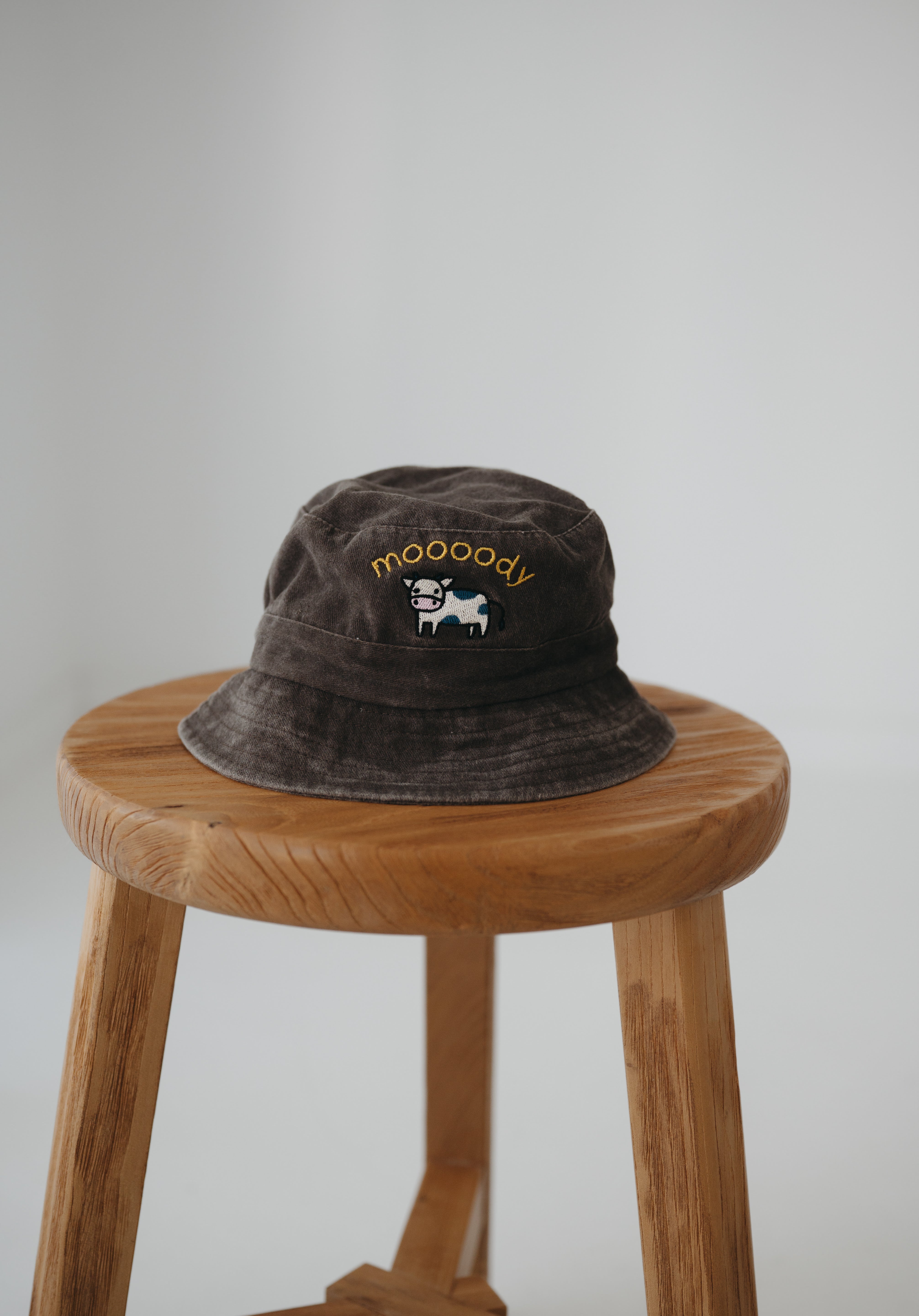A brown washed cotton daycare bucket hat with "moooody" embroidered in yellow with an embroidered cow, displayed on a wooden stool against a light grey background.