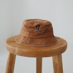 A brown washed cotton daycare bucket hat with "any bites yet" embroidered in blue with a an embroidered fish, displayed on a wooden stool against a light grey background.