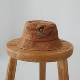 A brown washed cotton daycare bucket hat with "any bites yet" embroidered in blue with a an embroidered fish, displayed on a wooden stool against a light grey background.