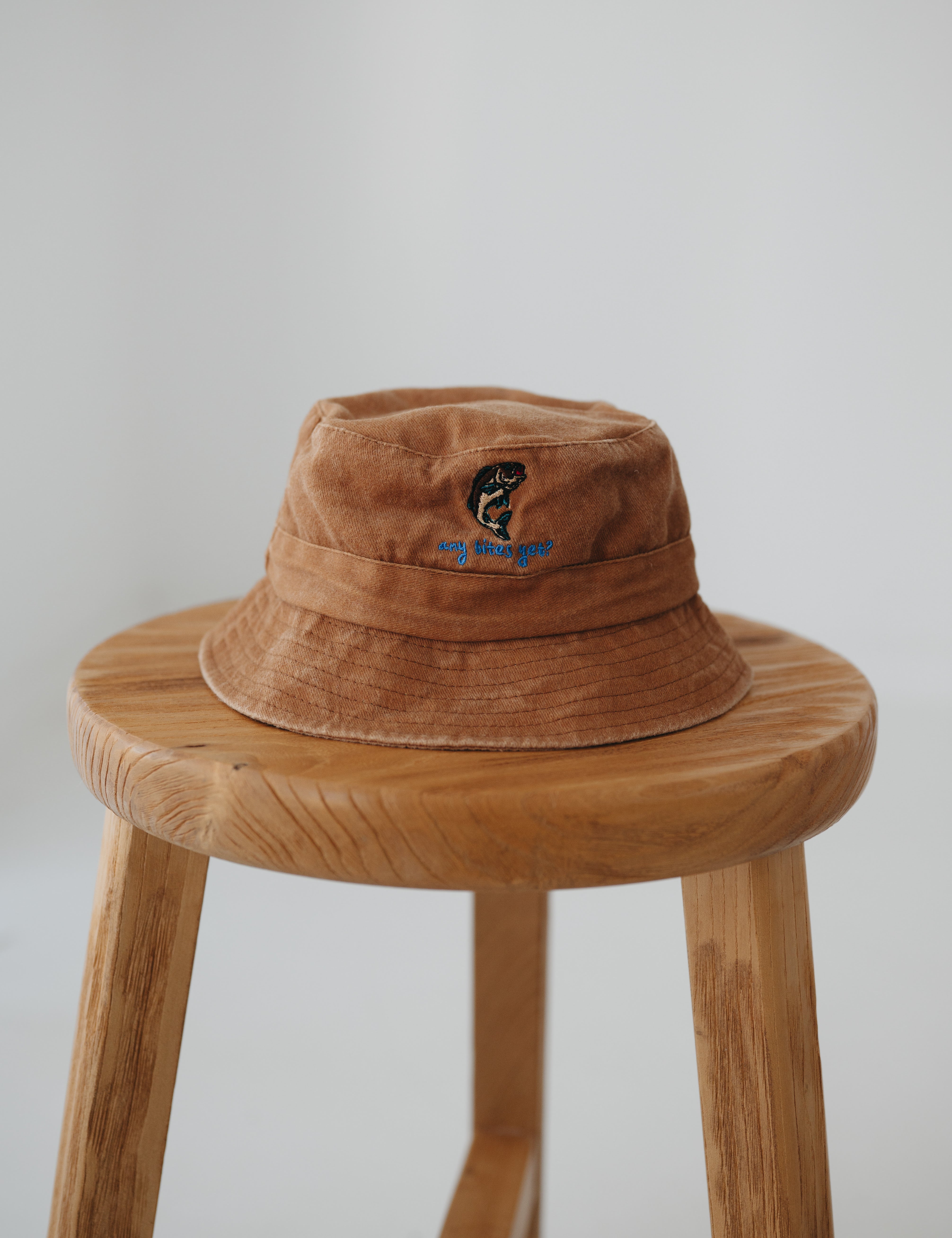 A brown washed cotton daycare bucket hat with "any bites yet" embroidered in blue with a an embroidered fish, displayed on a wooden stool against a light grey background.