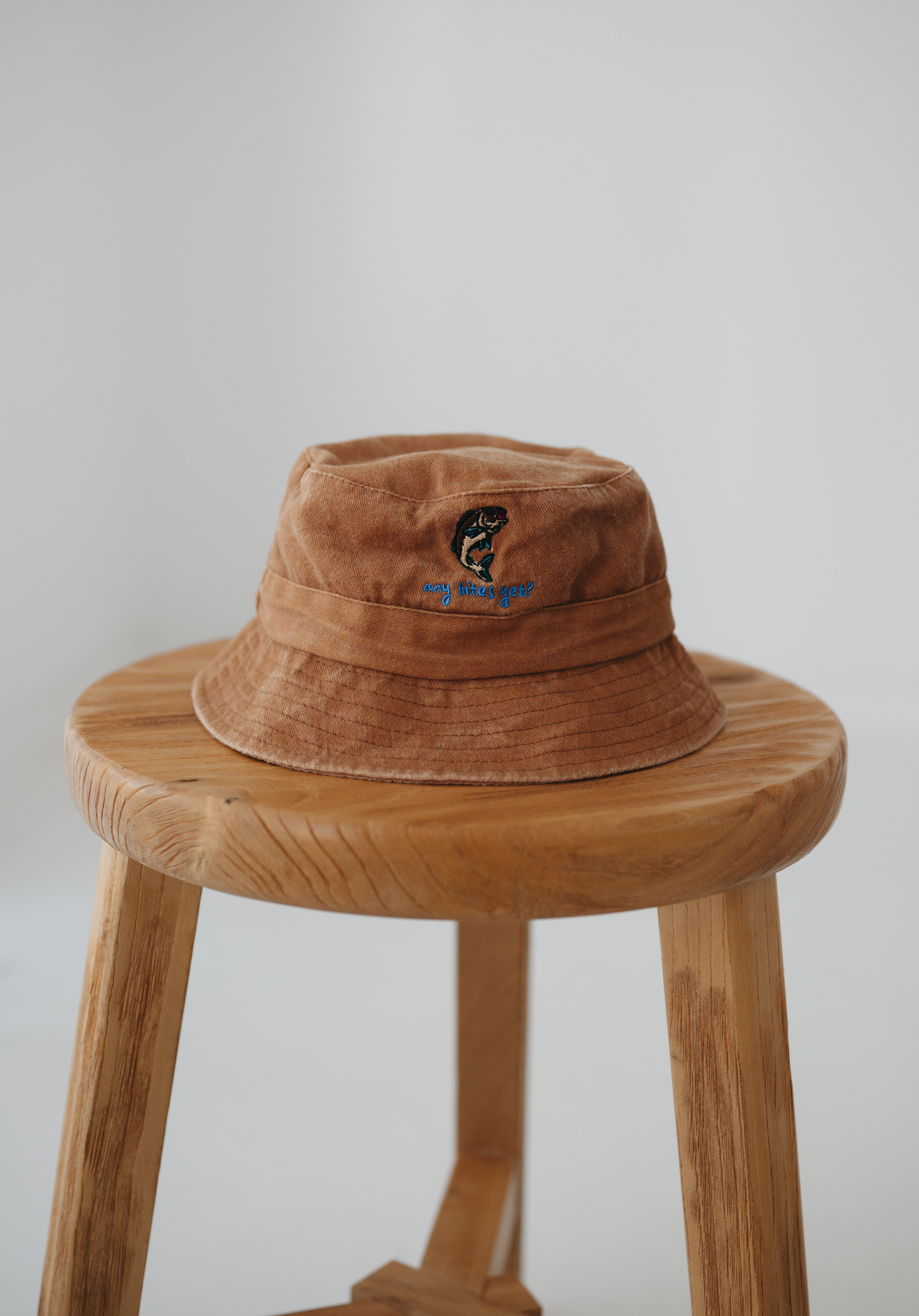 A brown washed cotton daycare bucket hat with "any bites yet" embroidered in blue with a an embroidered fish, displayed on a wooden stool against a light grey background.