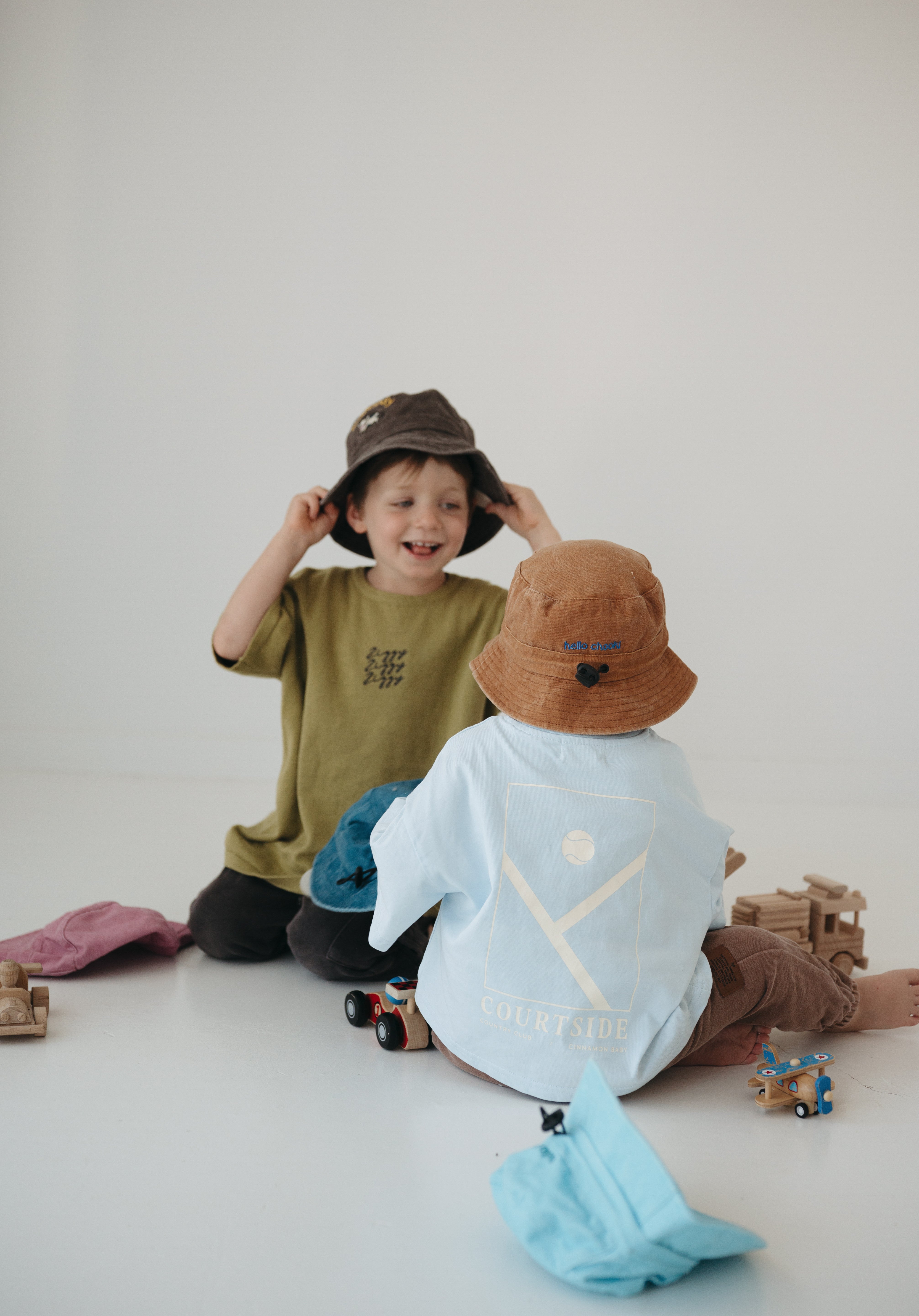 Two toddlers sitting on a white floor wearing a light brown washed cotton daycare bucket and a dark brown washed cotton daycare bucket with oversized t-shirts in a minimalist studio setting. One child wears an olive green t-shirt and a dark brown hat, while the other wears a light blue t-shirt  a light brown hat.
