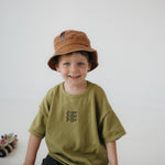 A toddler smiling at the camera kneeling on a white floor, wearing an olive green oversized T-shirt, dark pants, and a washed cotton brown daycare bucket hat.