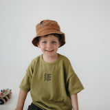 A toddler smiling at the camera kneeling on a white floor, wearing an olive green oversized T-shirt, dark pants, and a washed cotton brown daycare bucket hat.