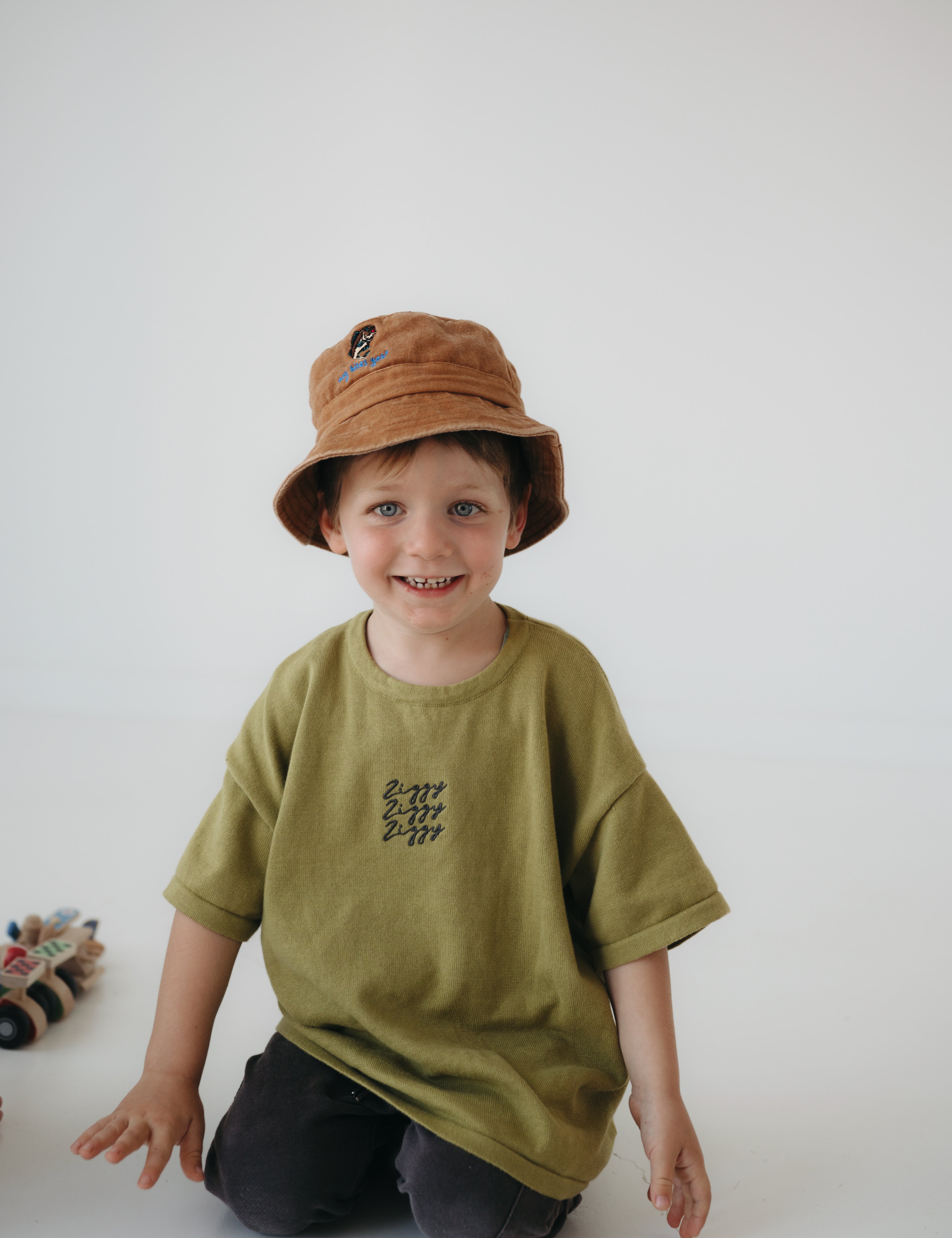 A toddler smiling at the camera kneeling on a white floor, wearing an olive green oversized T-shirt, dark pants, and a washed cotton brown daycare bucket hat.
