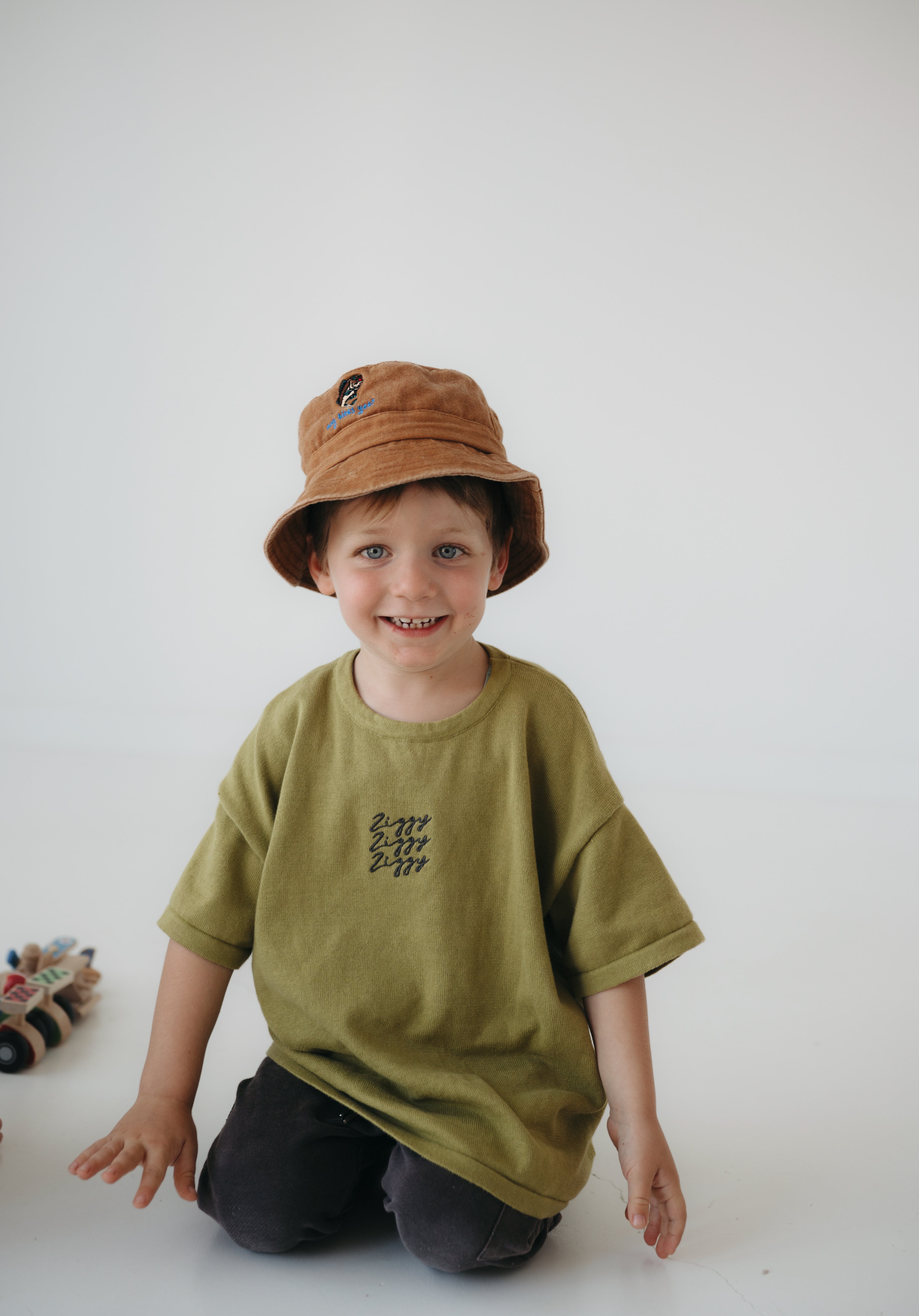 A toddler smiling at the camera kneeling on a white floor, wearing an olive green oversized T-shirt, dark pants, and a washed cotton brown daycare bucket hat.