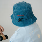 A dark blue washed cotton toddler daycare bucket hat with "hello chooki" embroidered in a red text on the back, displayed on a round wooden stool against a white background.