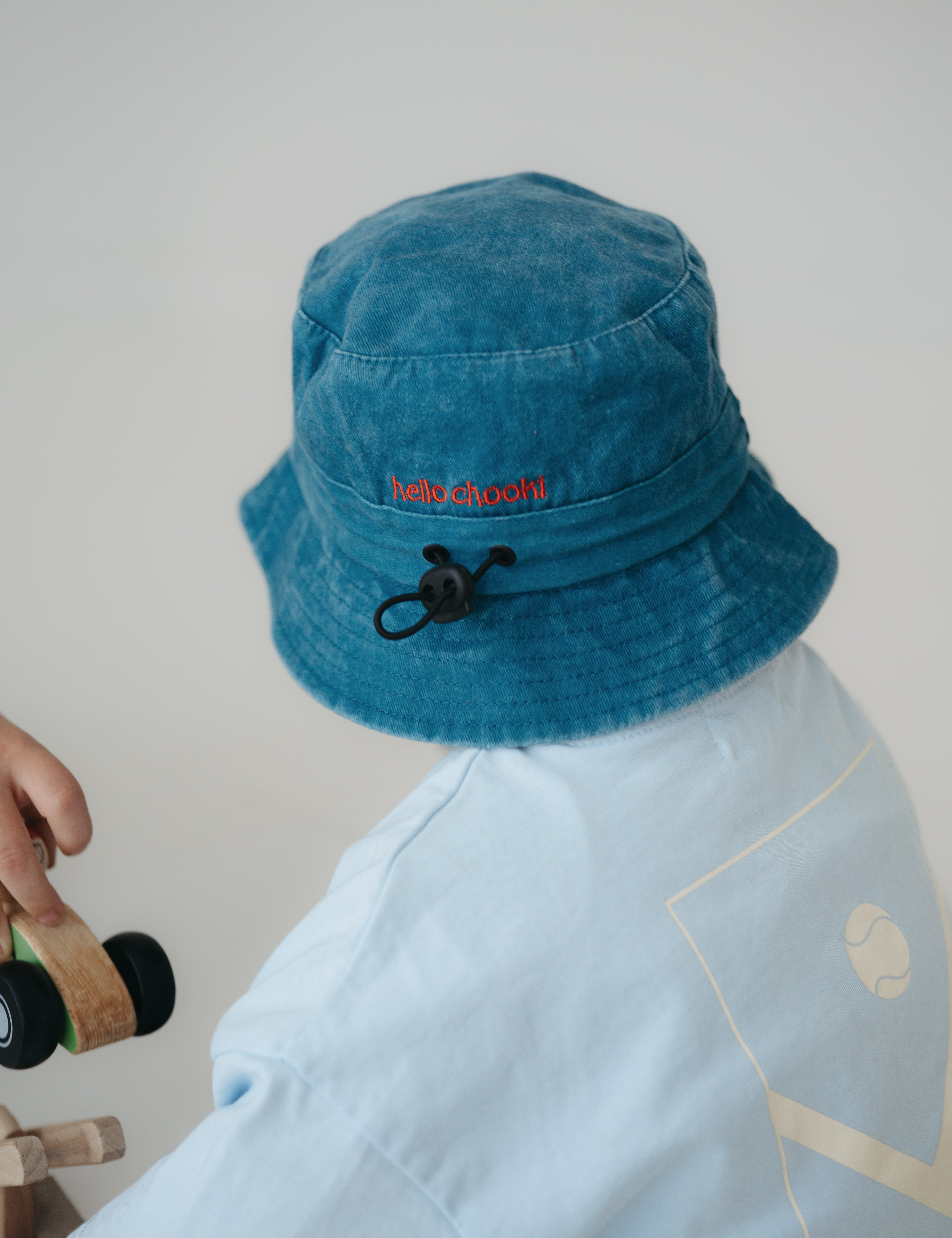 A dark blue washed cotton toddler daycare bucket hat with "hello chooki" embroidered in a red text on the back, displayed on a round wooden stool against a white background.