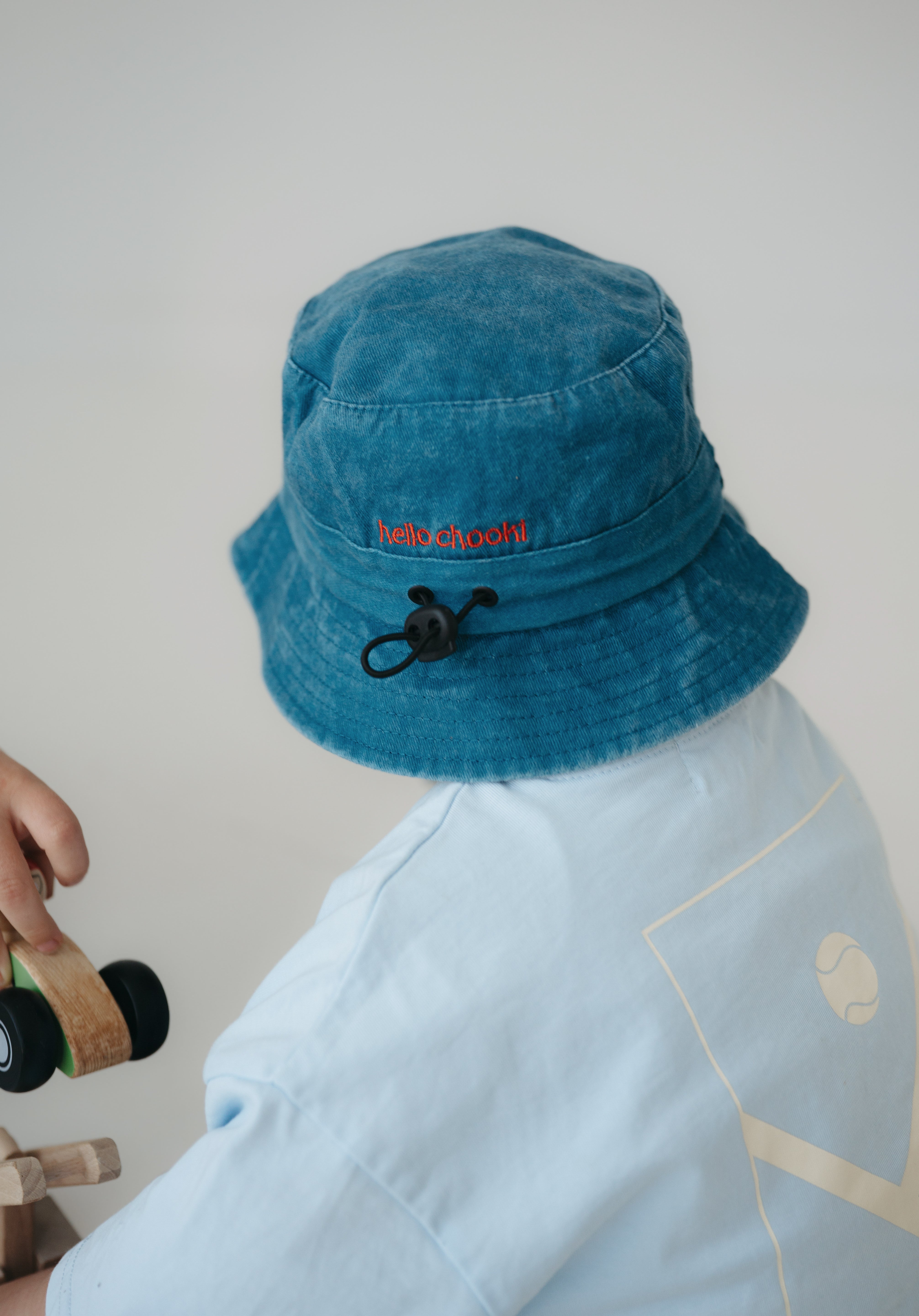 A dark blue washed cotton toddler daycare bucket hat with "hello chooki" embroidered in a red text on the back, displayed on a round wooden stool against a white background.