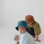 Two toddlers in oversized t-shirts pose on a seamless white studio floor. The front child wears a teal washed cotton daycare bucket hat and light blue shirt, while the back child wears a brown washed cotton daycare bucket hat.