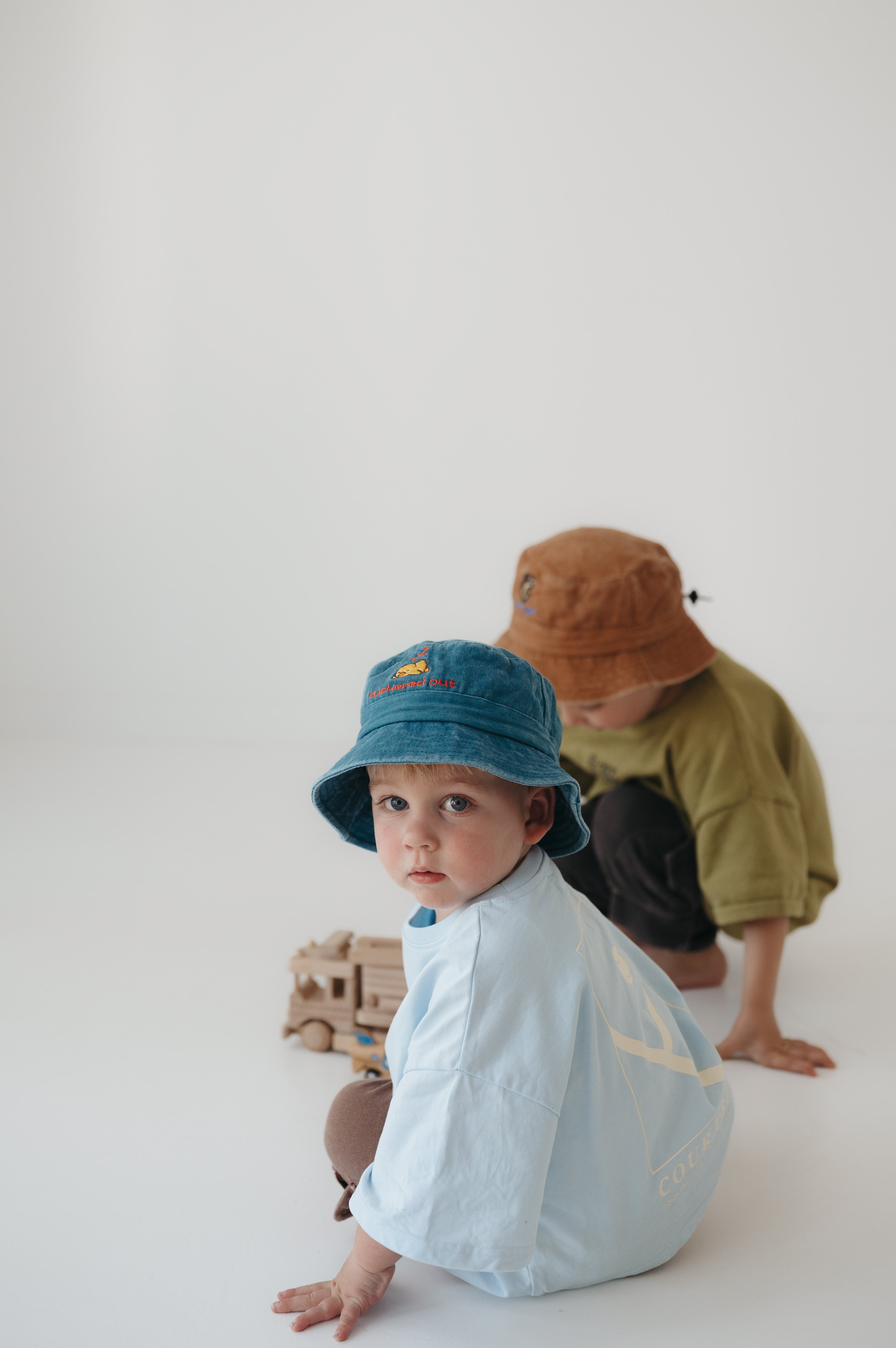 Two toddlers in oversized t-shirts pose on a seamless white studio floor. The front child wears a teal washed cotton daycare bucket hat and light blue shirt, while the back child wears a brown washed cotton daycare bucket hat.