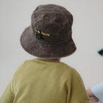 A toddler viewed from behind wearing a dark brown washed cotton bucket hat with "hello chooki" in yellow embroidered text and an olive green shirt, set against a plain light grey wall.