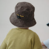 A toddler viewed from behind wearing a dark brown washed cotton bucket hat with "hello chooki" in yellow embroidered text and an olive green shirt, set against a plain light grey wall.