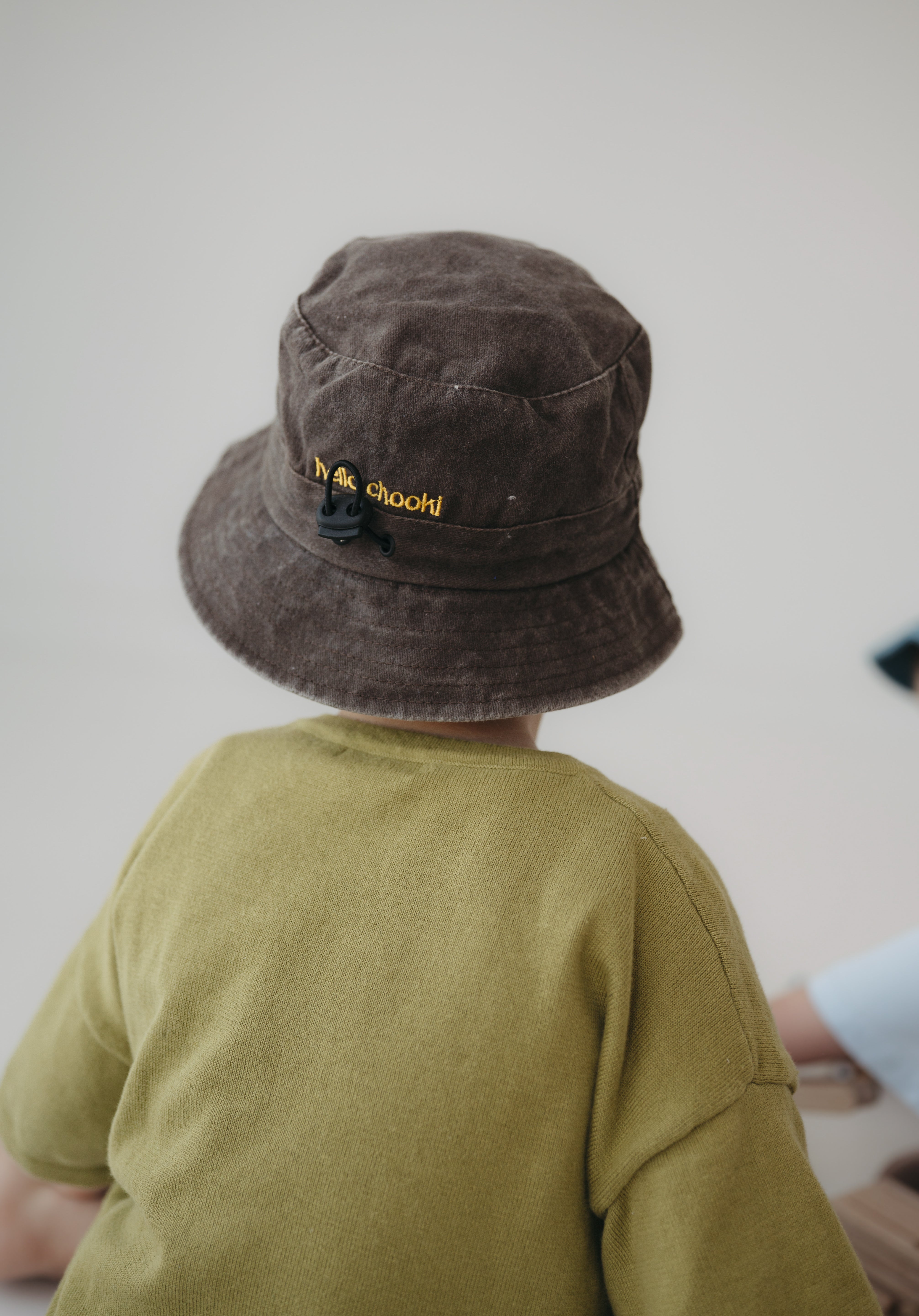 A toddler viewed from behind wearing a dark brown washed cotton bucket hat with "hello chooki" in yellow embroidered text and an olive green shirt, set against a plain light grey wall.
