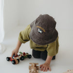 A toddler wearing a dark brown washed cotton bucket hat with 'moody' embroidered on the front in yellow, kneeling on a white floor while playing with wooden toy cars.