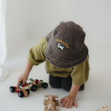 A toddler wearing a dark brown washed cotton bucket hat with 'moody' embroidered on the front in yellow, kneeling on a white floor while playing with wooden toy cars.
