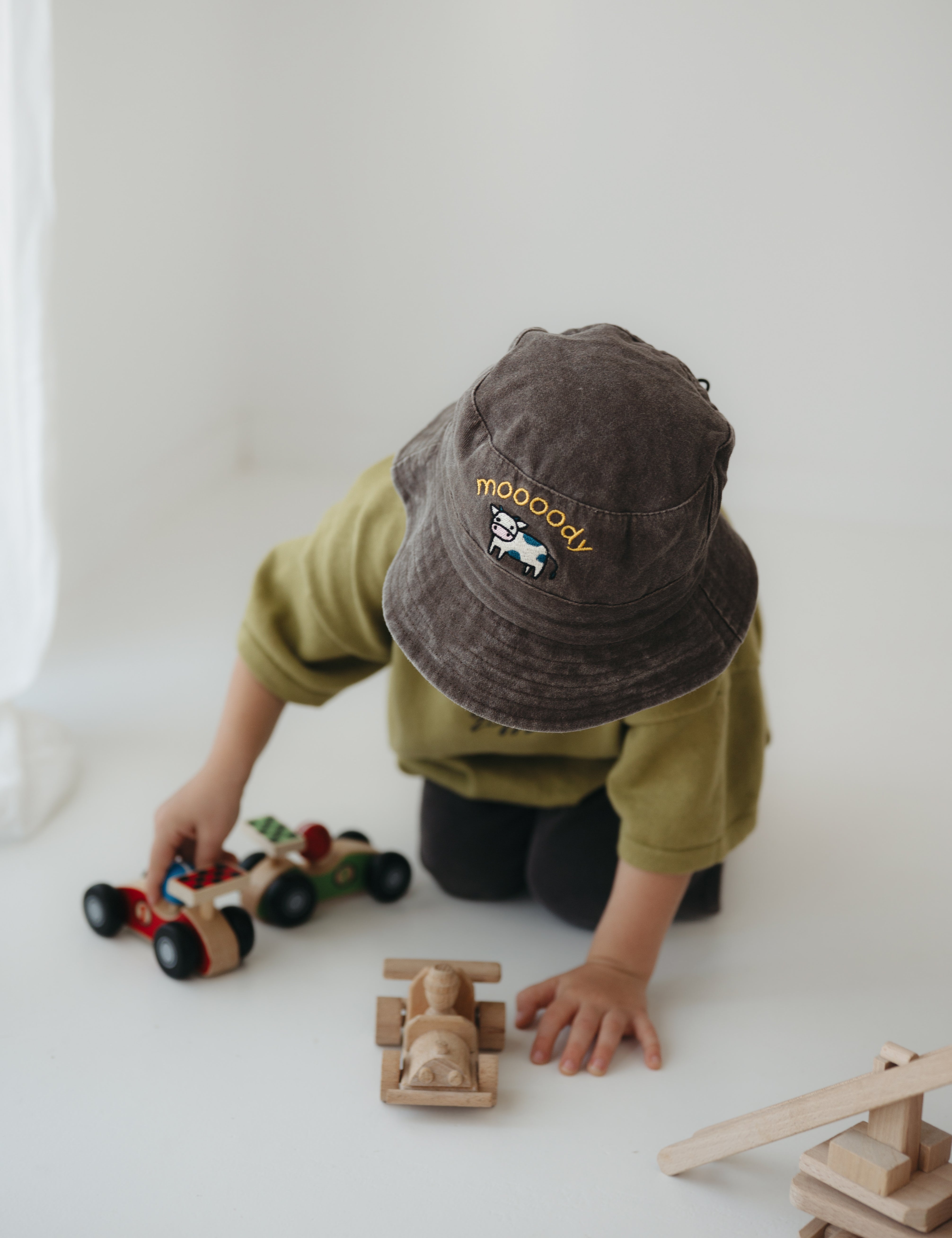A toddler wearing a dark brown washed cotton bucket hat with 'moody' embroidered on the front in yellow, kneeling on a white floor while playing with wooden toy cars.