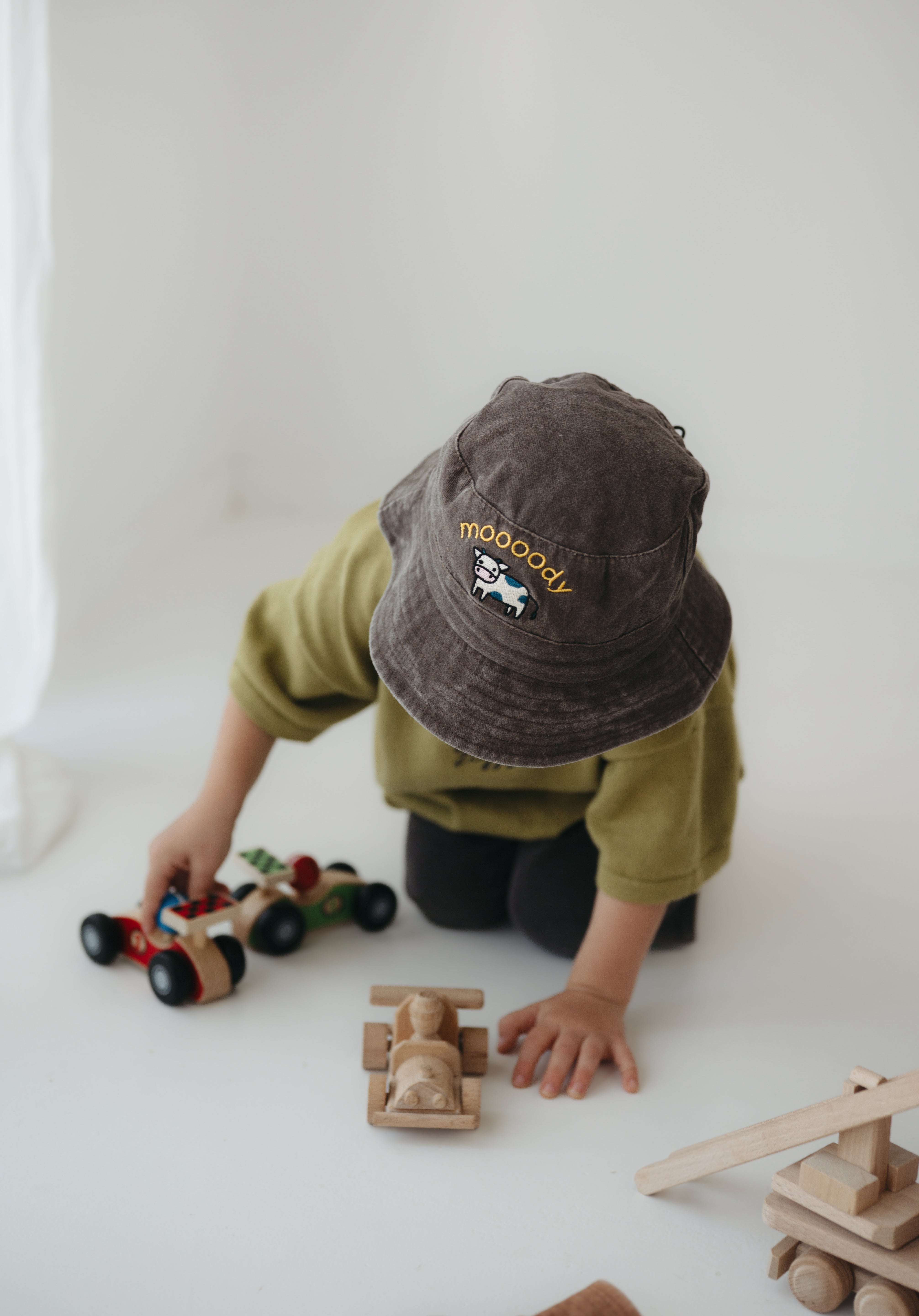A toddler wearing a dark brown washed cotton bucket hat with 'moody' embroidered on the front in yellow, kneeling on a white floor while playing with wooden toy cars.