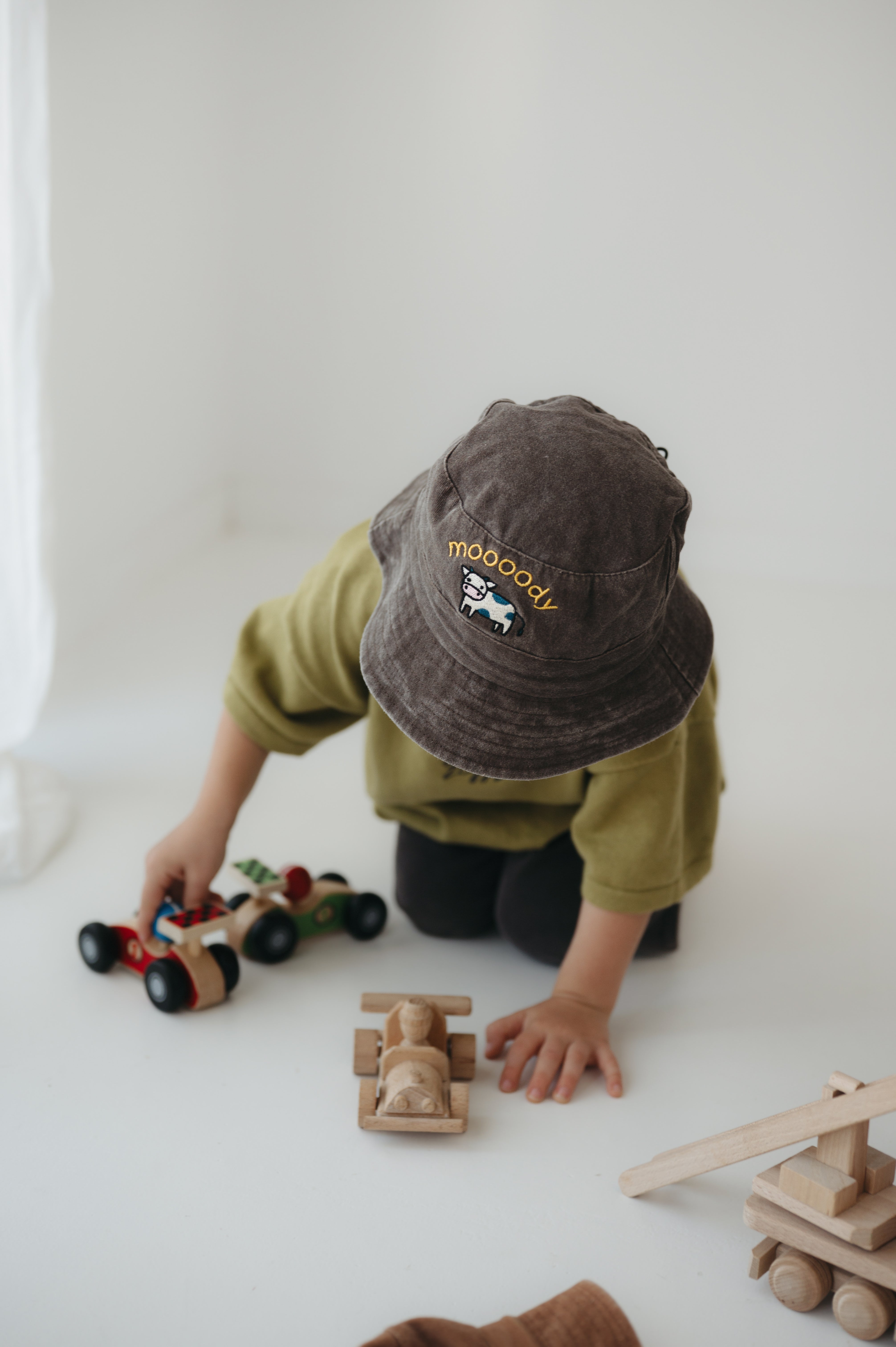 A toddler wearing a dark brown washed cotton bucket hat with 'moody' embroidered on the front in yellow, kneeling on a white floor while playing with wooden toy cars.