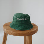A dark green washed cotton toddler daycare bucket hat with "daycare day" embroidered in a very soft pink text on the front, displayed on a round wooden stool against a white background.