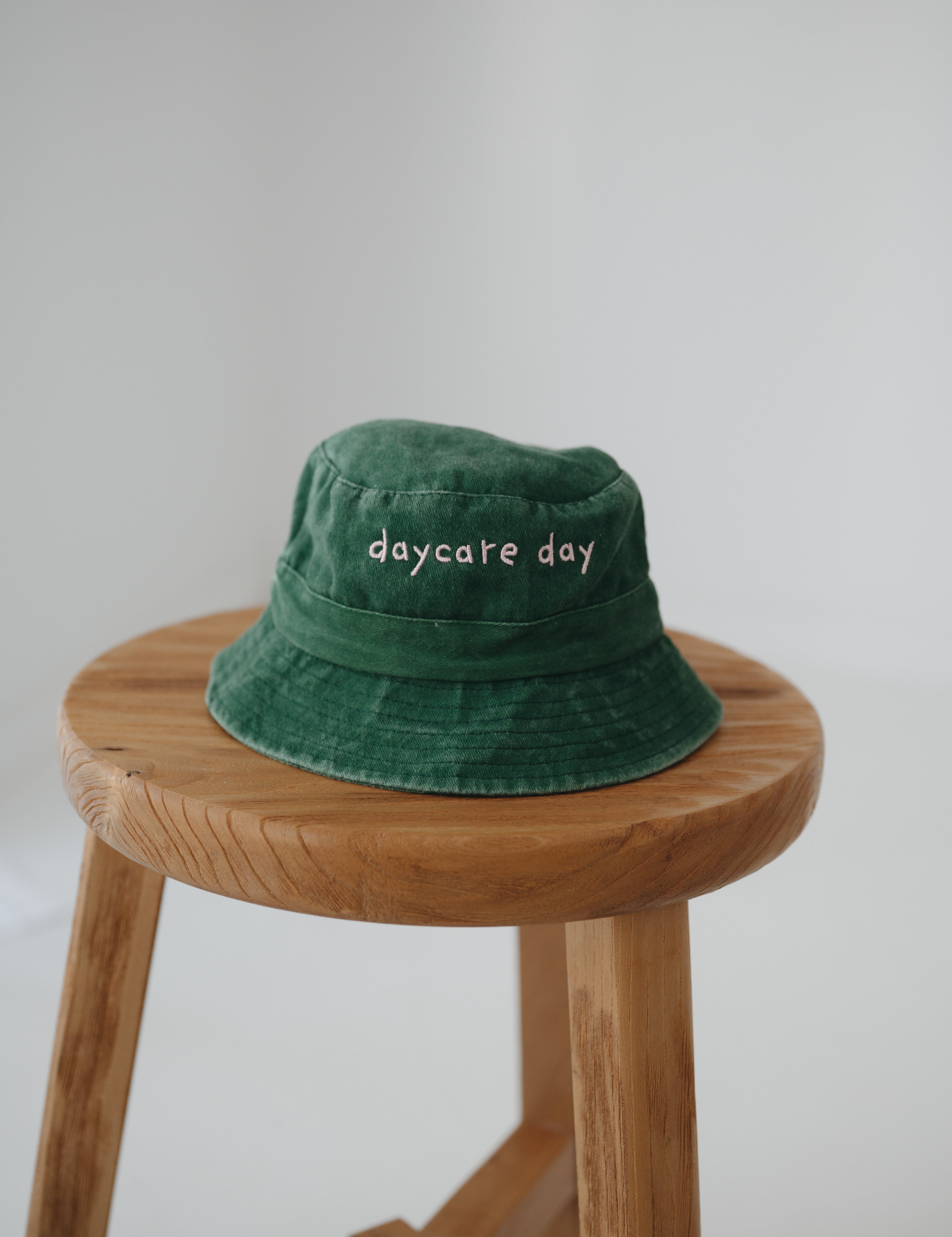 A dark green washed cotton toddler daycare bucket hat with "daycare day" embroidered in a very soft pink text on the front, displayed on a round wooden stool against a white background.