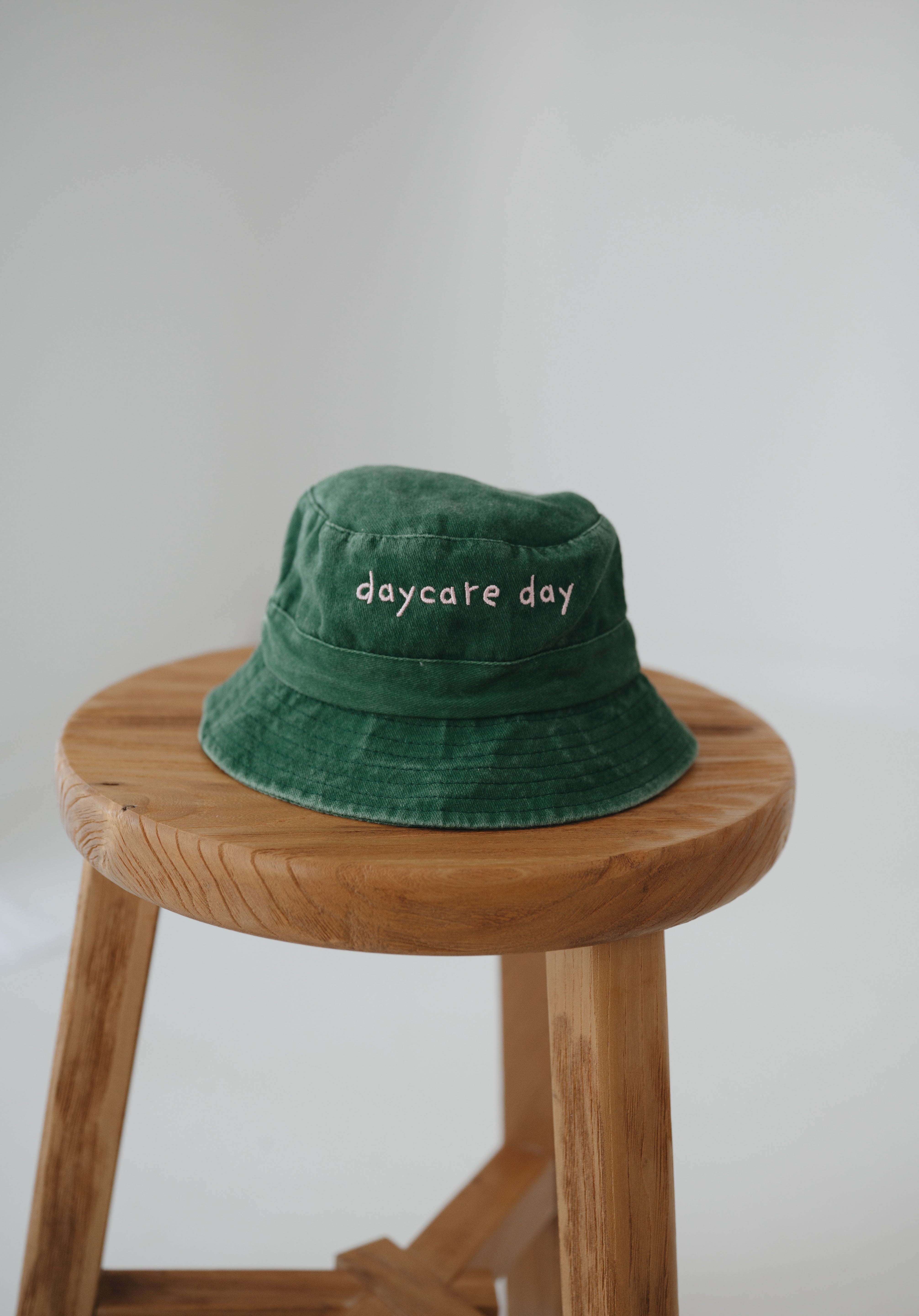A dark green washed cotton toddler daycare bucket hat with "daycare day" embroidered in a very soft pink text on the front, displayed on a round wooden stool against a white background.