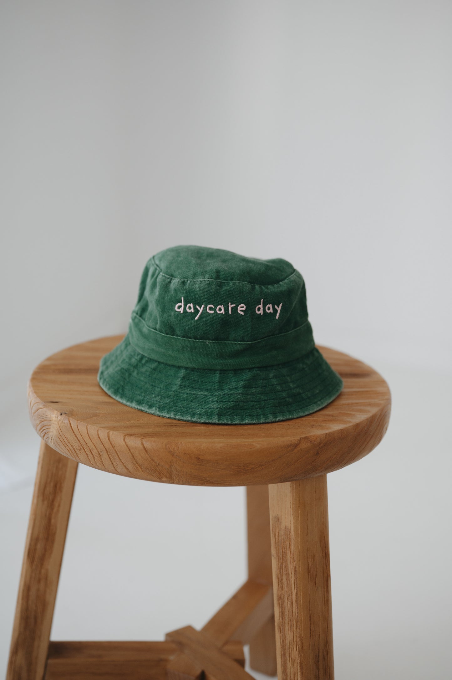 A dark green washed cotton toddler daycare bucket hat with "daycare day" embroidered in a very soft pink text on the front, displayed on a round wooden stool against a white background.