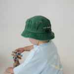A toddler wearing a washed cotton green daycare bucket hat with 'hello chooki' embroidered on the back and a light blue t-shirt, seen from behind while playing with wooden truck and plane against a plain light background.