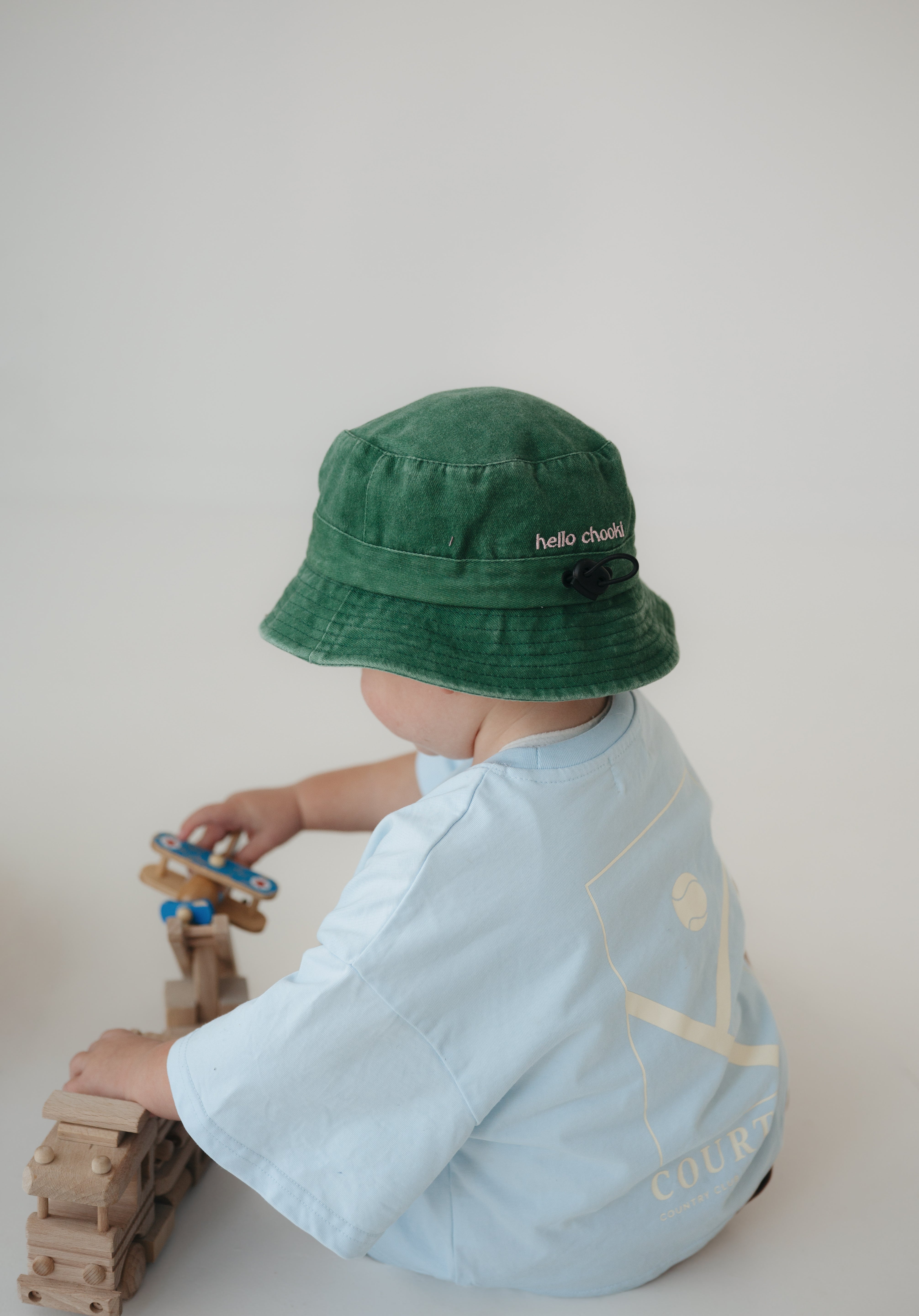 A toddler wearing a washed cotton green daycare bucket hat with 'hello chooki' embroidered on the back and a light blue t-shirt, seen from behind while playing with wooden truck and plane against a plain light background.