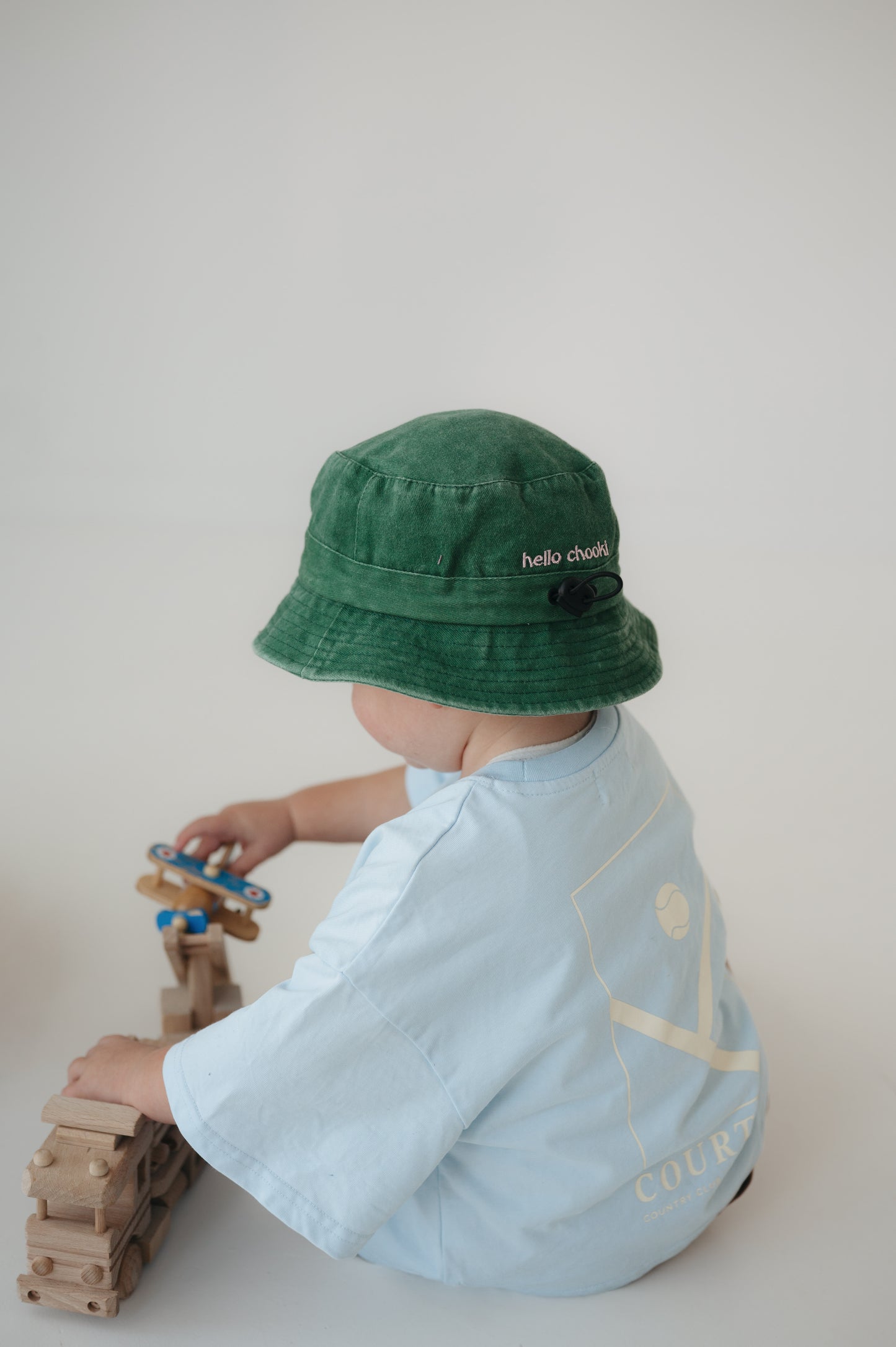 A toddler wearing a washed cotton green daycare bucket hat with 'hello chooki' embroidered on the back and a light blue t-shirt, seen from behind while playing with wooden truck and plane against a plain light background.