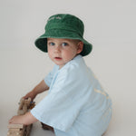 A toddler wearing a green washed cotton daycare bucket hat and a light blue short-sleeve shirt sits on a light grey studio floor, holding a wooden toy train.