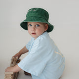 A toddler wearing a green washed cotton daycare bucket hat and a light blue short-sleeve shirt sits on a light grey studio floor, holding a wooden toy train.