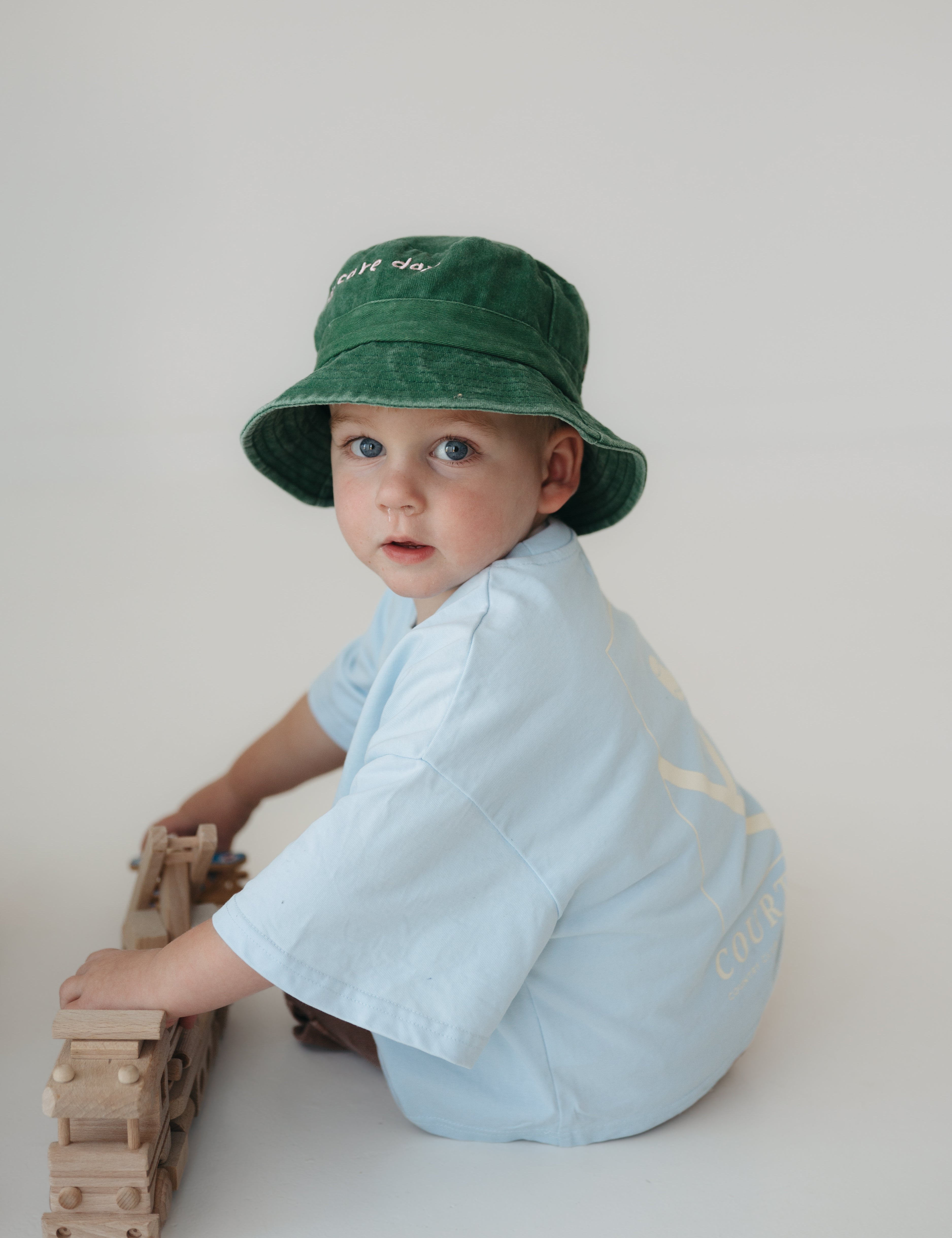 A toddler wearing a green washed cotton daycare bucket hat and a light blue short-sleeve shirt sits on a light grey studio floor, holding a wooden toy train.