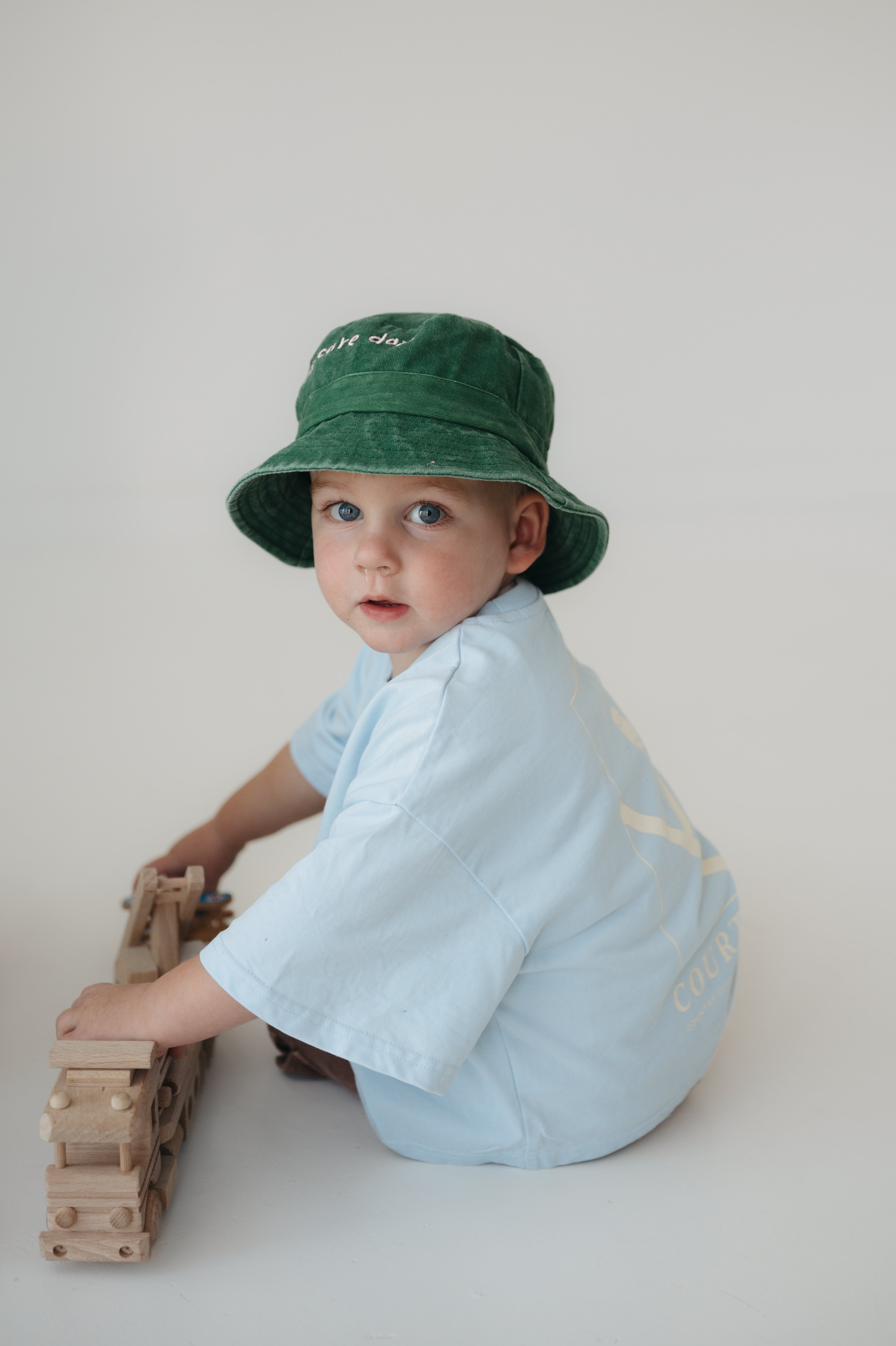 A toddler wearing a green washed cotton daycare bucket hat and a light blue short-sleeve shirt sits on a light grey studio floor, holding a wooden toy train.