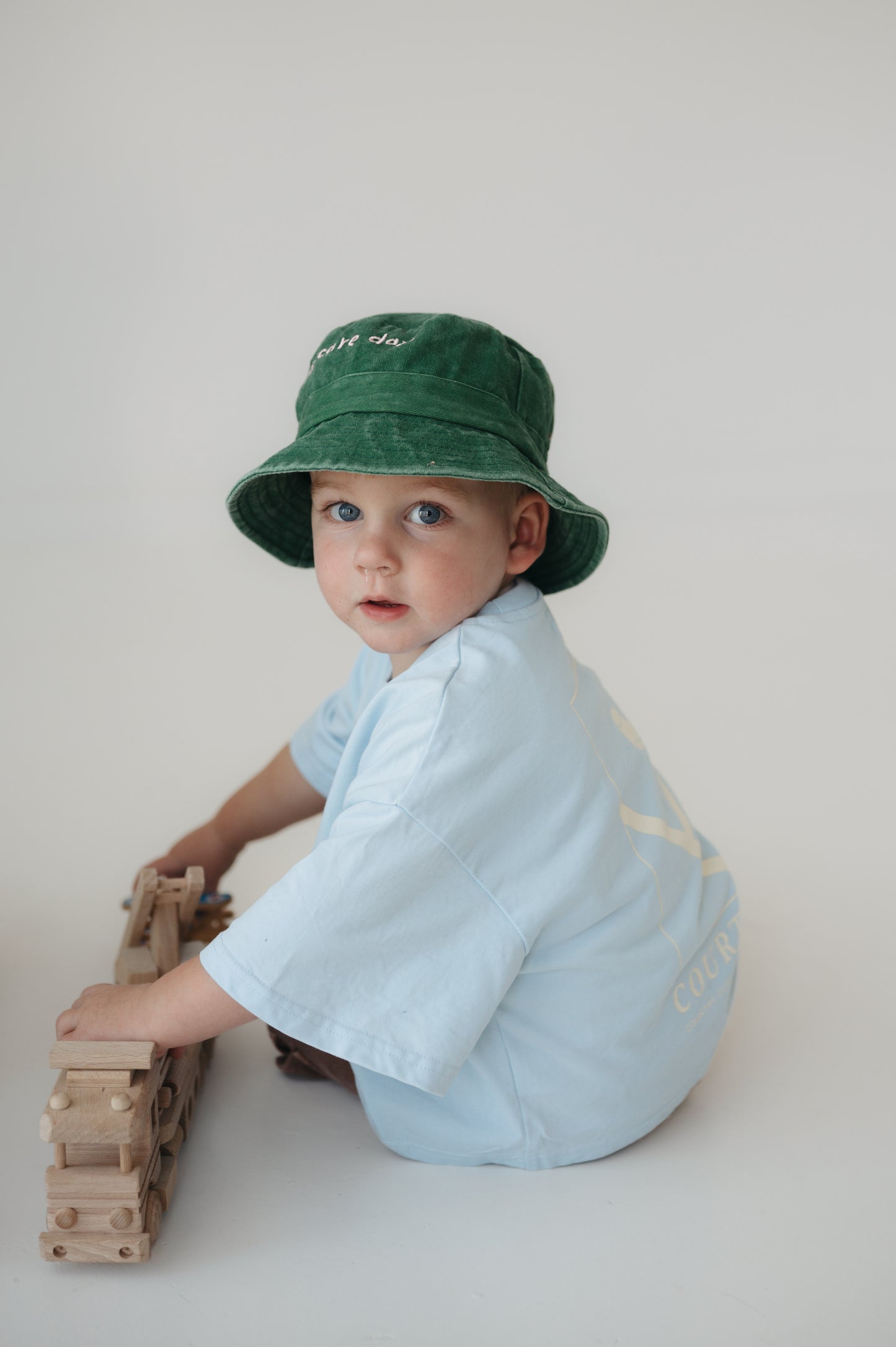 A toddler wearing a green washed cotton daycare bucket hat and a light blue short-sleeve shirt sits on a light grey studio floor, holding a wooden toy train.
