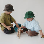 Two toddlers sitting on a white floor playing with wooden blocks and a toy car, wearing a dark brown washed cotton daycare bucket hat and a green washed cotton daycare bucket hat with casual t-shirts in a bright, minimalist room.