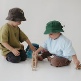 Two toddlers sitting on a white floor playing with wooden blocks and a toy car, wearing a dark brown washed cotton daycare bucket hat and a green washed cotton daycare bucket hat with casual t-shirts in a bright, minimalist room.